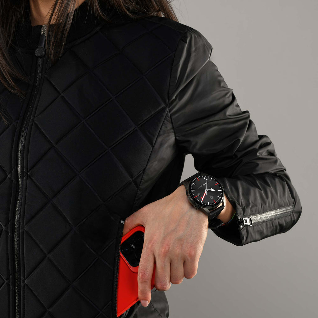 Giacca trapuntata nera e telefono rosso in mano, al polso l'orologio rotondo Momo Design Zero